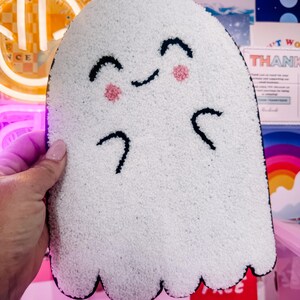 Op de afbeelding: Een witte, spookvormige pluche met een lachend gezicht. De geest heeft zwart geborduurde ogen, wenkbrauwen en armen, met roze blosjes op de wangen. De pluche heeft een zwarte rand en een getextureerd oppervlak.