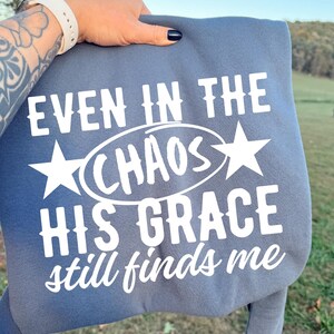 May include: A blue-grey sweatshirt with the text "EVEN IN THE CHAOS HIS GRACE still finds me" in white. The word "CHAOS" is inside an oval, with a star on each side. The sweatshirt is held up by a person.
