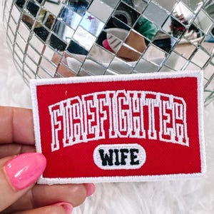Può includere: Una toppa rettangolare rossa con testo ricamato bianco che recita "FIREFIGHTER" sopra "WIFE" in un ovale bianco. La toppa ha un bordo bianco. Una palla da discoteca è sullo sfondo.