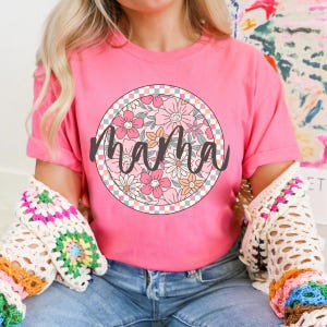 Puede incluir: Camiseta rosa con un diseño floral en un círculo con un borde a cuadros. La palabra "mama" está escrita en letras cursivas negras sobre el diseño floral.
