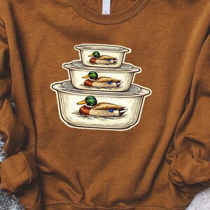 May include: A brown sweatshirt featuring a graphic of three stacked, vintage-style containers, each with a duck illustration. The ducks have green heads, brown bodies, and blue wing accents. The containers are white with gray lids.