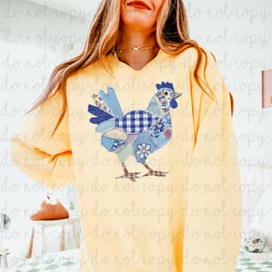 May include: A yellow sweatshirt featuring a patchwork chicken design in shades of blue, white, and floral patterns. The chicken has a blue comb and beak, with a checkered pattern on its body. The sweatshirt is a solid yellow color.