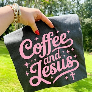 May include: Dark gray t-shirt with the words "Coffee and Jesus" in pink script. The design includes stars and a cross. The shirt is held up against a green background.
