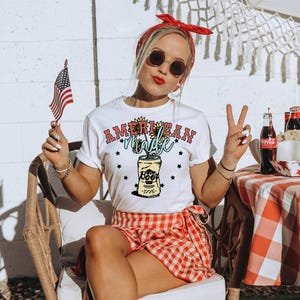 Puede incluir: Camiseta blanca con un gráfico de una lata de cerveza con girasoles y el texto "AMERICAN made" y estrellas. La lata tiene el texto "Beer" y "1976".