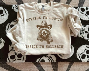 Peut inclure: Un sweat-shirt beige avec un raton laveur brun portant un chapeau de cowboy et le texte "Outside I'm Hootin' Inside I'm Hollerin'"