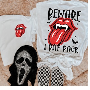 May include: White t-shirts with Halloween-themed graphics. One shirt features a red tongue with fangs, and the text "BEWARE I BITE BACK." Another shirt has a red tongue graphic. A Scream mask and checkered shoes are also in the image.