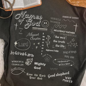 Pode incluir: Camiseta cinza escuro com as palavras "Nomes de Deus" e outros termos religiosos em branco. Uma Bíblia e uma pulseira dourada são visíveis no fundo. Uma bandana amarela também está no fundo.