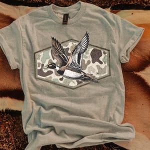 以下が含まれることがあります： 飛んでいるアヒルの詳細なイラストが描かれた、明るい灰色のTシャツ。アヒルは六角形のフレーム内の迷彩模様を背景にしています。シャツは柔らかい素材でできており、半袖です。