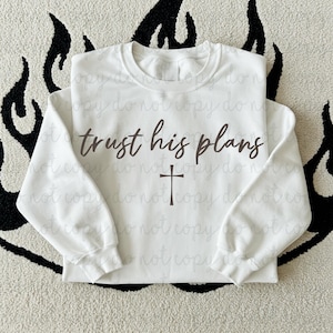 May include: White crew neck sweatshirt with the text "trust his plans" in a brown script font, and a brown cross below the text. The sweatshirt is displayed on a patterned surface.