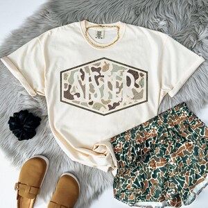 May include: Cream-colored t-shirt with the word "SISTER" in a camouflage design. Matching camouflage shorts are next to the shirt. A gold chain necklace and brown sandals complete the look.