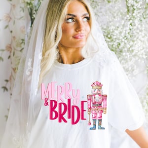 Puede incluir: Camiseta blanca con el texto "Merry & Bride" en letras rosas y una ilustración de cascanueces rosa y dorado.