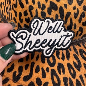 May include: A leopard print hat with a black and white patch that says "Well Sheeit". The hat has a brown and black spotted pattern. The patch is white with black cursive lettering and a black border.