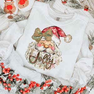 May include: White Christmas sweatshirt featuring a watercolour illustration of Santa Claus wearing a red hat with white polka dots and a gold bow. The word "Jolly" is written in gold glitter lettering.