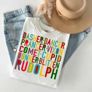 May include: White t-shirt with a colorful graphic design featuring the names of Santa's reindeer: Dasher, Dancer, Prancer, Vixon, Comet, Cupid, Donner, Blitzen, and Rudolph.
