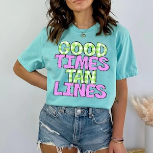 May include: A light blue t-shirt with the words "GOOD TIMES TAN LINES" in a playful font. The words are in green and pink, with a checkered pattern. The shirt is a comfortable, casual style.