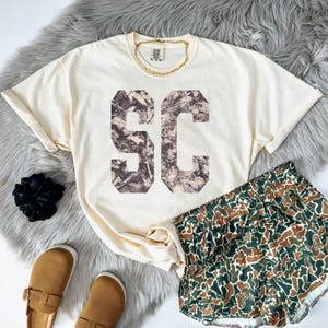 May include: Cream-colored t-shirt with the letters "SC" in a camouflage pattern. The shirt is paired with matching camouflage shorts, brown sandals, and a gold chain necklace. A black scrunchie is also visible.