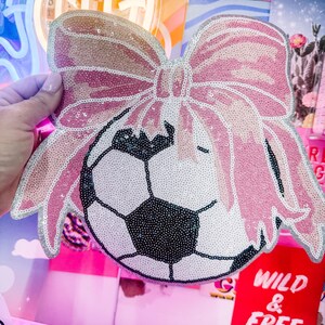 May include: A decorative soccer ball with a pink sequined bow. The soccer ball is black and white, and the bow is a light pink with silver trim. The item is likely a decorative accessory or a novelty item.