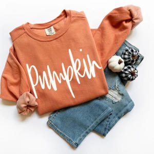 Könnte beinhalten: Ein orangefarbenes Sweatshirt mit dem Wort "Pumpkin" in weißer Schrift. Das Sweatshirt wird mit hellblauen Jeans und dekorativen Kürbissen kombiniert. Die Ärmel sind umgeschlagen und zeigen einen helleren Orangeton.
