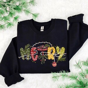 Puede incluir: Sudadera negra con la palabra "MERRY" en letras festivas y coloridas. Las letras están decoradas con lunares, luces navideñas y adornos. La sudadera tiene mangas largas y cuello redondo.