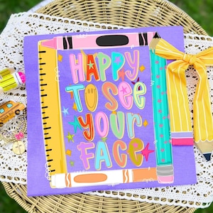 May include: A purple t-shirt featuring a graphic of a yellow ruler, a pink crayon, an orange marker, and a teal pencil surrounding the text HAPPY TO SEE YOUR FACE in colorful letters, with a yellow pencil-patterned fabric bow attached to the side.