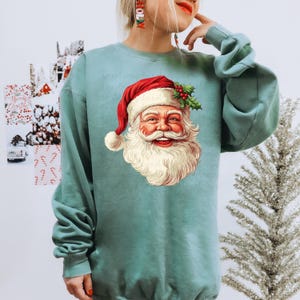 Puede incluir: Una sudadera de color verde azulado con un gráfico de la cara de Papá Noel. Papá Noel lleva un gorro rojo con un pompón blanco y una barba blanca. Acebo y bayas adornan el gorro. La sudadera tiene mangas largas y cuello redondo.