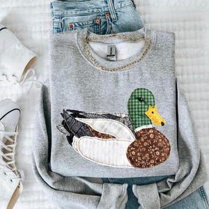 May include: A gray sweatshirt featuring a patchwork duck design with green, brown, and white patterned details. The sweatshirt is paired with blue jeans, a gold chain necklace, and white sneakers.