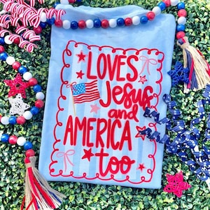 May include: Light blue t-shirt with red text that reads "LOVES Jesus and AMERICA too" with an American flag graphic. The shirt is decorated with red stars and bows. The background includes red, white, and blue decorative beads and ribbons.