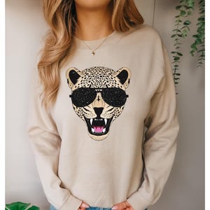 May include: A beige sweatshirt with a graphic print of a cheetah wearing sunglasses. The cheetah has black spots and a pink nose.
