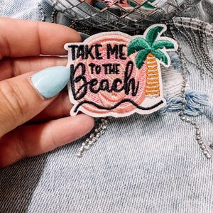 Puede incluir: Un parche bordado que presenta un fondo circular rosa con una palmera verde y amarilla. El texto "TAKE ME TO THE BEACH" está escrito en hilo negro. El parche tiene un borde blanco y una línea negra ondulada en la parte inferior con costuras.