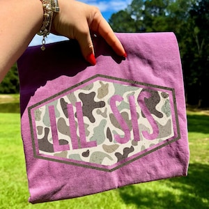 May include: A purple t-shirt with a camouflage design inside a hexagon shape. The design features the text "LIL SIS" in a pink font. The camouflage pattern includes shades of gray, brown, and green.
