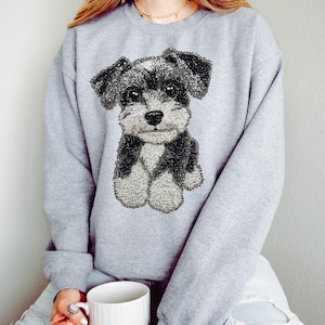 Puede incluir: Una sudadera gris con una ilustración detallada de un perro Schnauzer. El perro está representado en tonos de negro, blanco y gris, con una textura esponjosa. La sudadera es de color gris liso.
