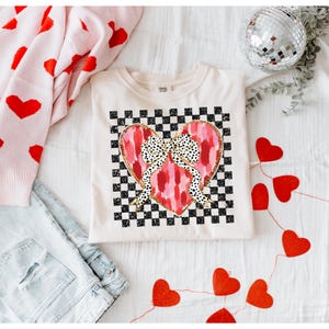 Peut inclure: Un t-shirt blanc avec un cœur rose et rouge sur un fond à carreaux noirs et blancs. Le cœur a un nœud doré avec des pois noirs.