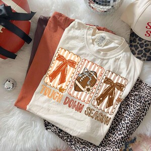 May include: Cream-colored t-shirt with a football graphic and the text "Tough Down Season." The shirt is paired with leopard print pants and other fall-themed items. The shirt has a crew neck and short sleeves.