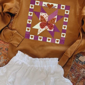 Puede incluir: Una sudadera marrón con un diseño inspirado en un edredón con un patrón de mariposa y corazones en tonos morado, blanco y rojo. Debajo se ve una falda blanca con volantes. El diseño está centrado en el pecho de la sudadera.