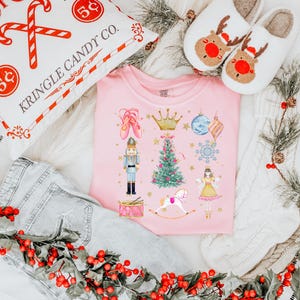May include: A pink t-shirt with a Christmas-themed design featuring a nutcracker, a Christmas tree, a ballerina, a rocking horse, a crown, a snowflake, a drum, and an ornament. The design is in a watercolor style.