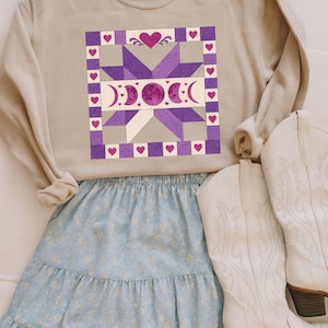 May include: A beige sweatshirt with a purple and gray quilt design featuring hearts and moon phases. Paired with a light blue floral skirt and white cowboy boots with brown heels. A casual, country-inspired outfit.