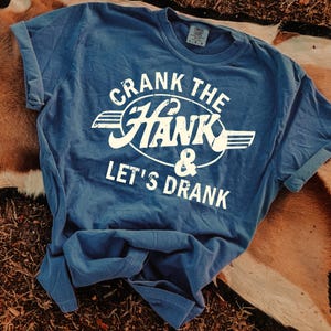 以下が含まれることがあります： 袖を折り返した青いTシャツ。白の掠れたフォントで「CRANK THE HANK & LET'S DRANK」の文字がプリントされています。茶色とベージュの表面に置かれています。
