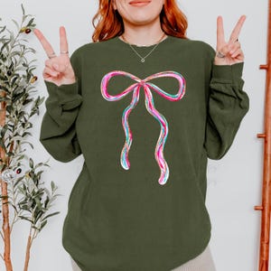 Puede incluir: Una sudadera verde oliva con un diseño de lazo colorido. El lazo está delineado en rosa y blanco, con detalles turquesa y dorados. La sudadera tiene mangas largas y cuello redondo.