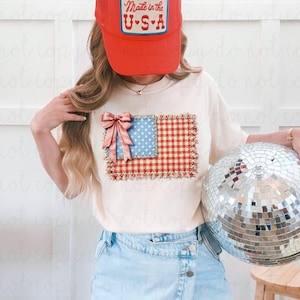 Könnte beinhalten: Ein cremefarbenes T-Shirt mit einem Patchwork-Design der amerikanischen Flagge, einer roten Schleife und dem Text "Made in the U.S.A." auf einer roten Baseballkappe. Das Flaggen-Design zeigt blaue Sterne und rotes Vichy. Eine silberne Discokugel wird gehalten.