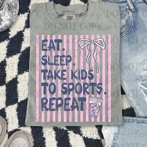 以下が含まれることがあります： ピンクと青で「EAT. SLEEP. TAKE KIDS TO SPORTS. REPEAT」と書かれたグレーのTシャツ。デザインにはリボンと氷の入った飲み物が含まれています。シャツは、ジーンズとスニーカーと一緒に市松模様のラグの上に表示されています。