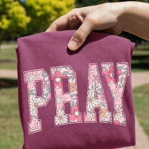 Pode incluir: Uma camiseta bordô com a palavra "PRAY" em letras grandes com estampas florais. As flores são em tons de rosa, branco e pêssego. A camiseta é exibida em um fundo verde desfocado.