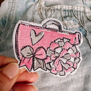 May include: Pink and white embroidered patch of a cheerleader megaphone with a bow and a heart. The patch is iron-on and can be used to decorate clothing, bags, or other items.