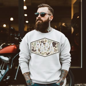 May include: White crew neck sweatshirt with a camouflage design in a hexagonal shape. The word "PAPA" is printed in white block letters within the camouflage pattern. The sweatshirt is worn by a person with a beard and sunglasses.