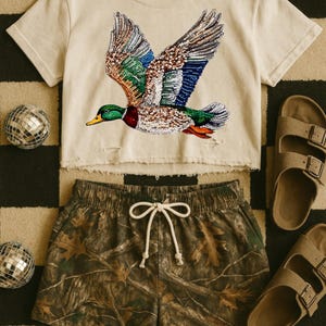 Op de afbeelding: Een crèmekleurige crop top met een pailletten eendenontwerp, gecombineerd met camouflage shorts en bruine sandalen. De eend heeft groene, blauwe en bruine pailletten. De shorts hebben een taille met trekkoord. De achtergrond is een zwart-wit dambordpatroon.