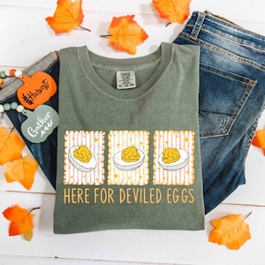 Könnte beinhalten: Salbeigrünes T-Shirt mit einer Grafik von drei gefüllten Eiern und dem Text "HERE FOR DEVILED EGGS" in Orange. Das Shirt wird mit blauen Jeans und herbstlichen Dekorationen präsentiert.