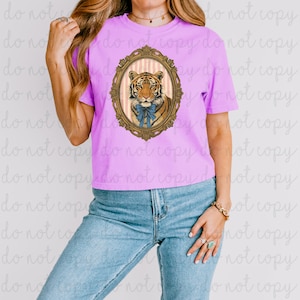 Puede incluir: Una camiseta de color lila con un retrato de tigre enmarcado. El tigre, adornado con un lazo azul, se encuentra sobre un fondo a rayas dentro de un marco dorado ornamentado. La camiseta se combina con vaqueros azules.