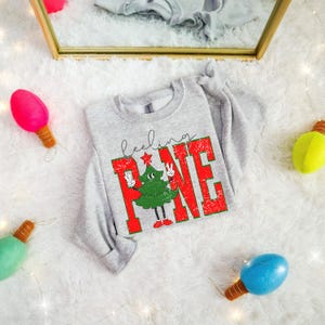 May include: A light grey sweatshirt with the words "feeling PINE" in red and green, featuring a cartoon Christmas tree. The sweatshirt is surrounded by colourful decorative lights.