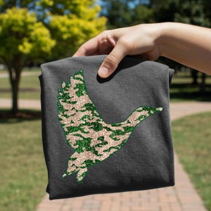 May include: A dark gray t-shirt featuring a glittery duck silhouette in a camouflage pattern of green and tan. The duck is in mid-flight, with its wings spread. The shirt is held up by a hand, showcasing the design.