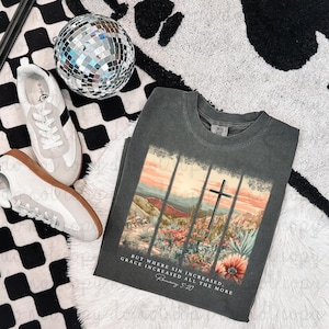 May include: A dark gray t-shirt featuring a landscape design with a cross and floral elements. The text "But where sin increased, grace increased all the more. Romans 5:20" is printed below the image. White sneakers and a disco ball are also visible.