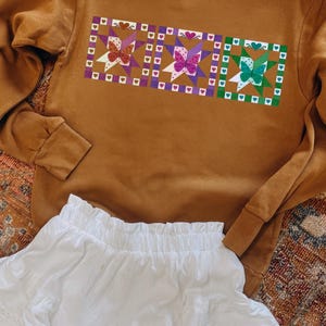 Puede incluir: Una sudadera de color mostaza con un diseño de mariposa colorido. El diseño presenta tres imágenes de mariposas en una fila, cada una dentro de un marco cuadrado con detalles de corazón y estrella. Debajo, una falda blanca con volantes.